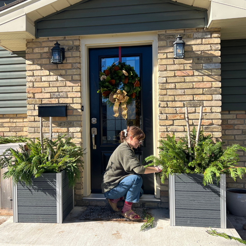 A person kneeling in front of a house between two large planter boxes, arranging winter greenery in a planter
