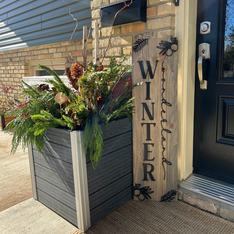 A large grey planter box arranged with christmas greenery outside a front door, next to a wooden sign that reads "winter wonderland"