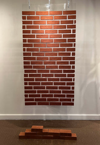A piece of plexiglass suspended from the ceiling is covred with thin-brick material so that there is space where the mortar should be in the brick. Shadowy text is visible on the wall behind the panel and can be read by peering through the brick.