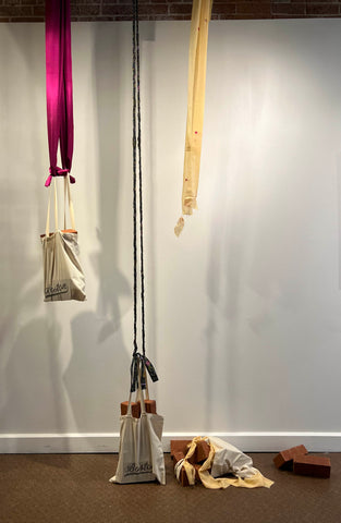Three canvas tourist bags with Boston in a script font. Each bag is full of red bricks. The left bag at head height is suspended by a magenta satin fabric. The center bag is suspended by a braided batik fabric, but it is so heavy it is suspended an inch from the floor. The right bag was suspended from a thin yellow sari fabric which has ripped under the weight - the bag has fallen to the floor and bricks have spilled out.