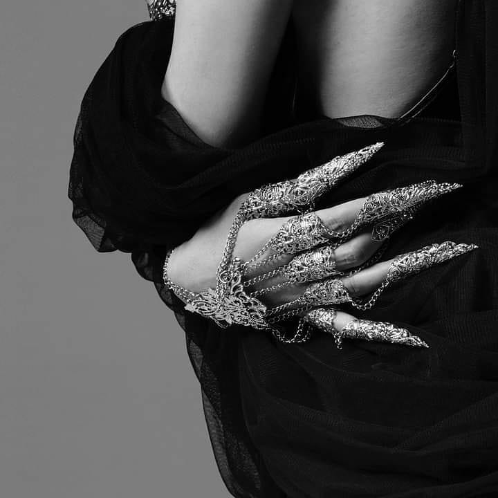 A grayscale image of a person's hand partially draped in a black garment, adorned with a striking silver filigree Metal Glove Diablo, showcasing elongated, ornate fingertip accents and interconnected chains, emphasizing a dramatic gothic aesthetic.