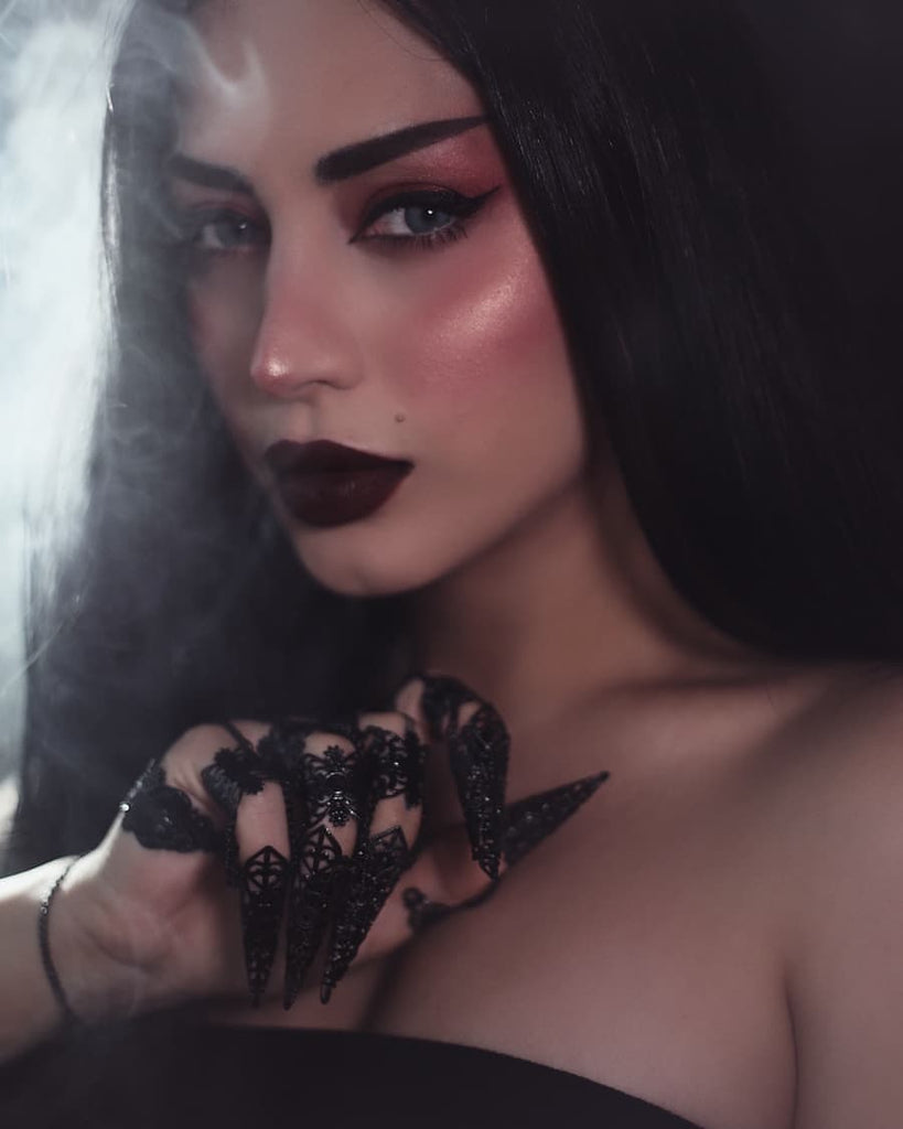 A gorgeous woman wearing dark cute makeup with strong black eyeliner wears an intricate hand jewel made of black filigrees that fully cover the hand. The claw rings are connected to the bracelet part with delicate black chains. It evokes a sense of refined goth style and dark fashion