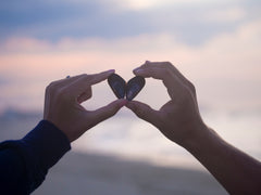 Two individuals are holding their hands up to the backdrop of the sky creating a heart by holding shells with their fingers