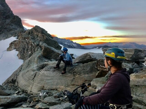 Two people wearing mountaineering helmets sitting on a mountain while the sun sets behind them