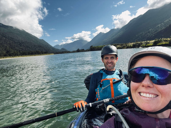 Man and woman smiling while packrafting down a river mountains in background