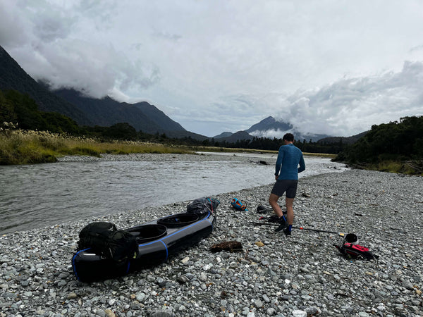Man standing by a river next to his packraft with mountains in the distance