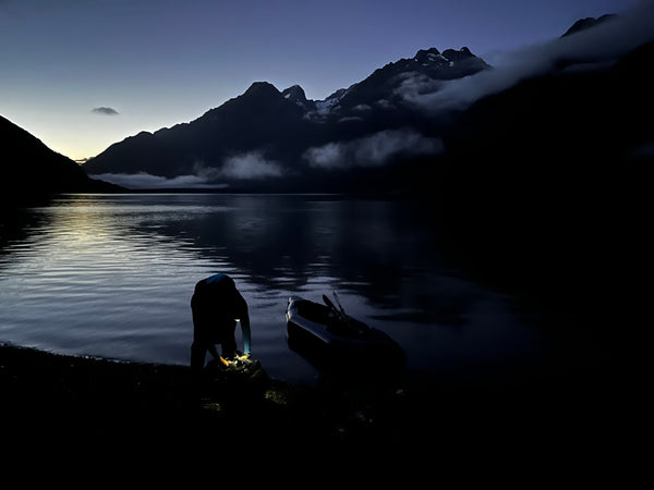 Person unpacking a packraft at dusk on the edge of a river with mountains in background