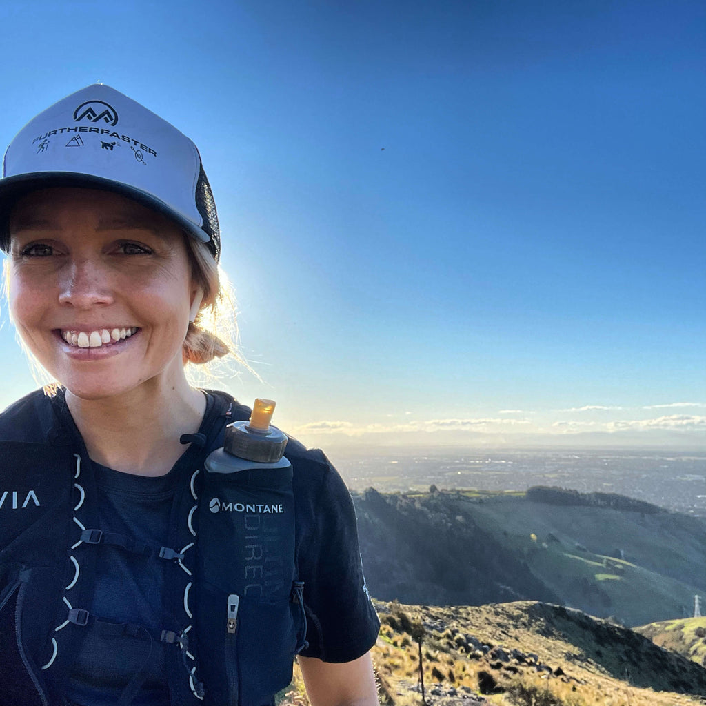 Woman smiling into the camera wearing a cap with mountain range in the distance.