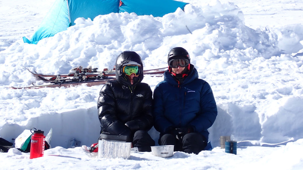 two people sitting in the snow wearing down jackets by a tent and skis.