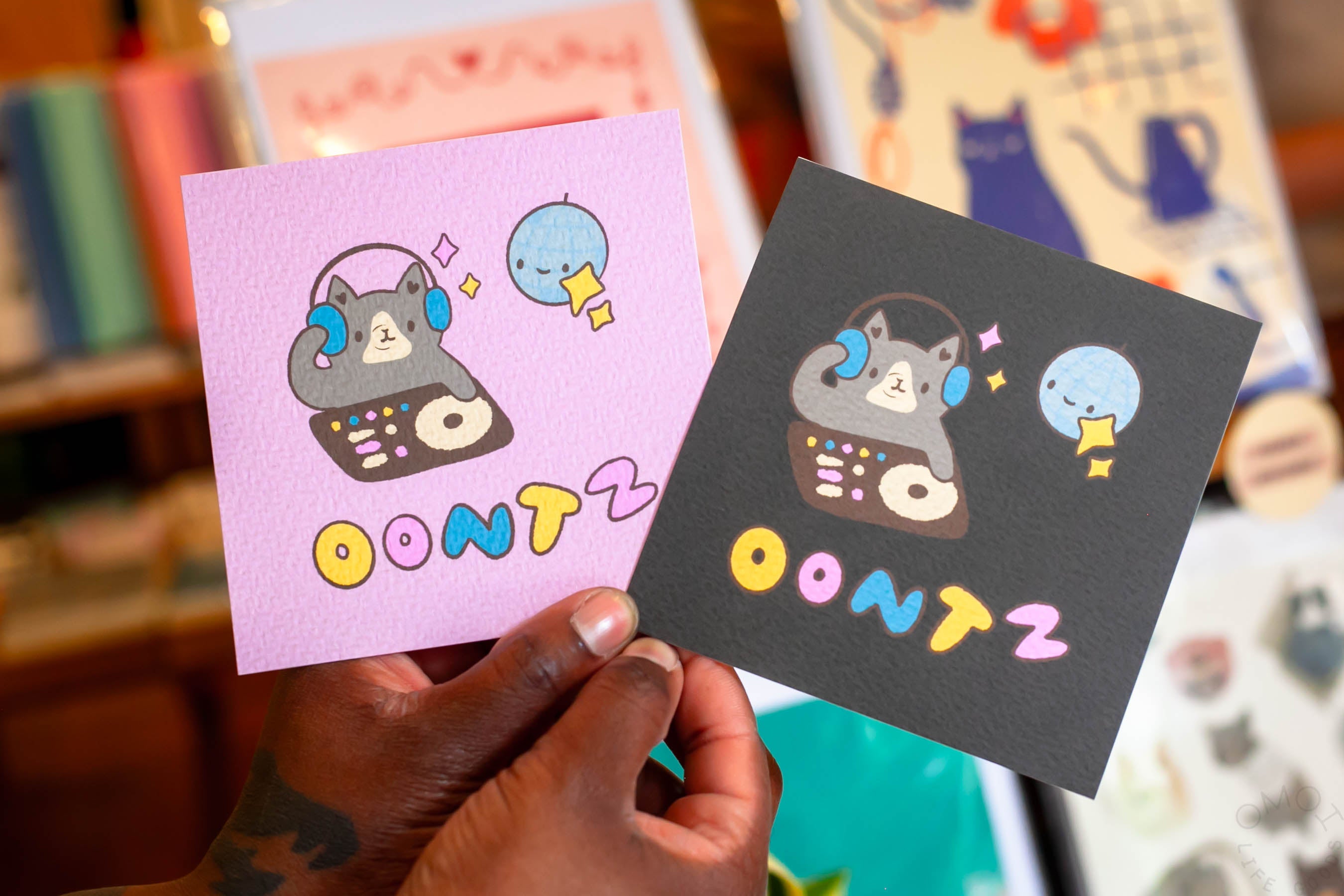 A closeup of square artwork, one on light purple background and another on black, both showing a cat DJ with the words OONTZ