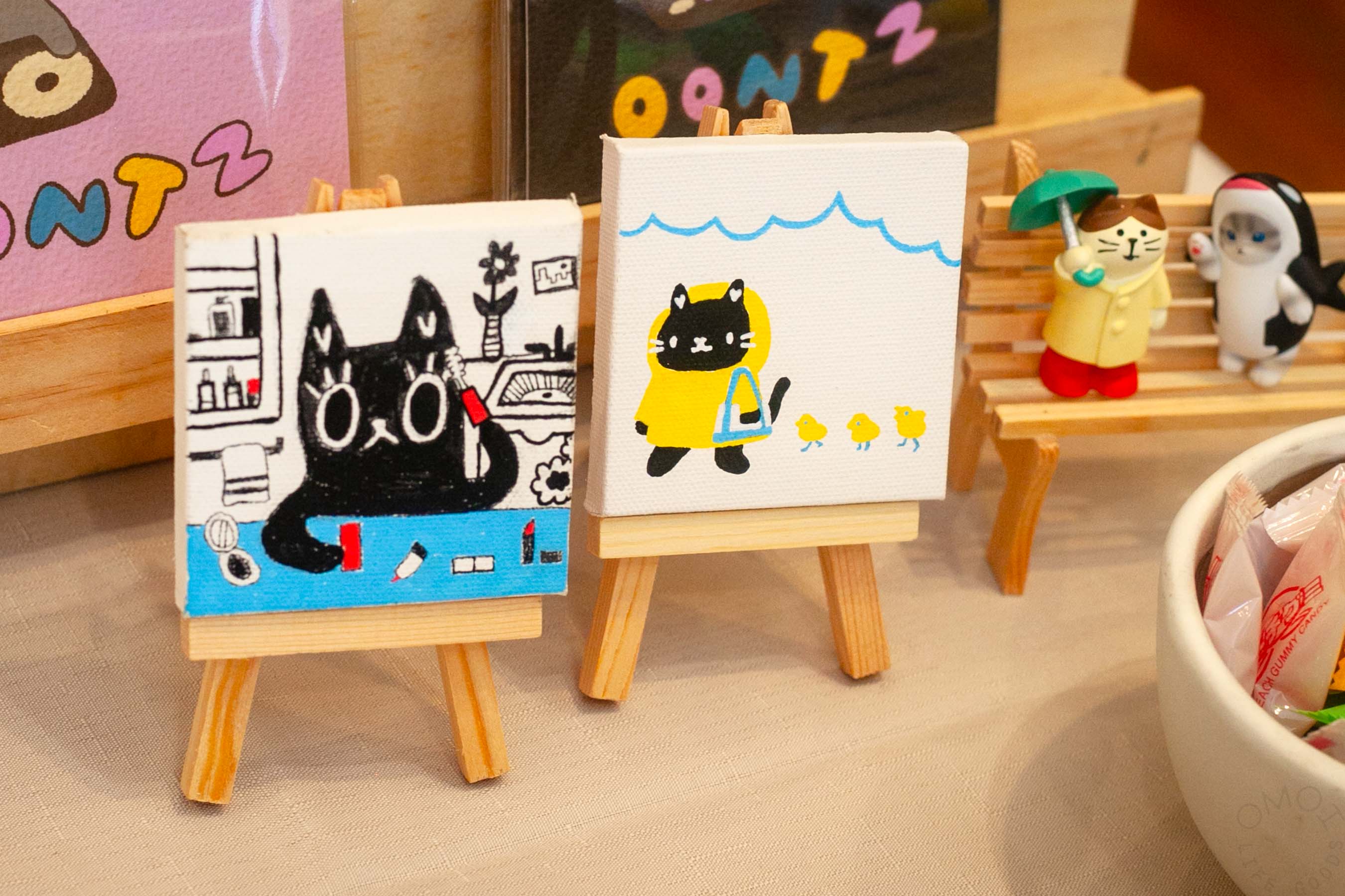 Tiny easels hold some Hannah Vy originals. Cute cat illustrations. One puts on makeup, another walks in the rain with some duckies.