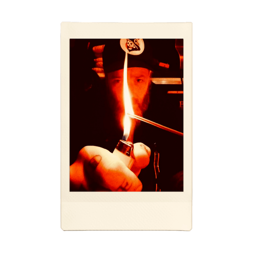 A scan of an Instax photo. A man in a trucker hat is holding a lighter to an incense stick before him. The flame grows high as it lights.