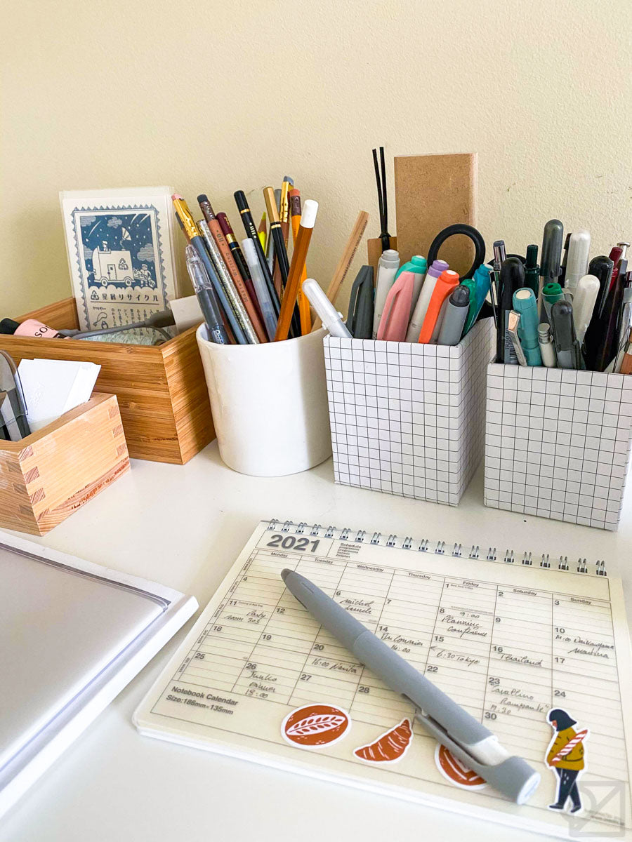 A white desk with a 2021 planner on it. There are square pen holders full of pens along the edge of the desk.