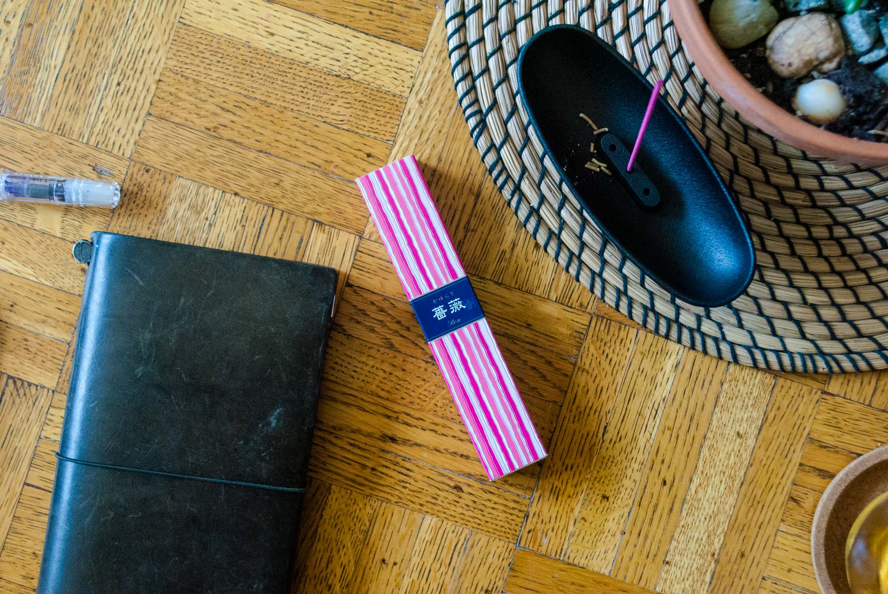 A rose-colored, slender rectangle package sits on a wood tabletop of bright orange undertones. Near the package is a cast iron incense burner shaped like a long, narrow oval. A rose-colored incense stick stands upright in its center.