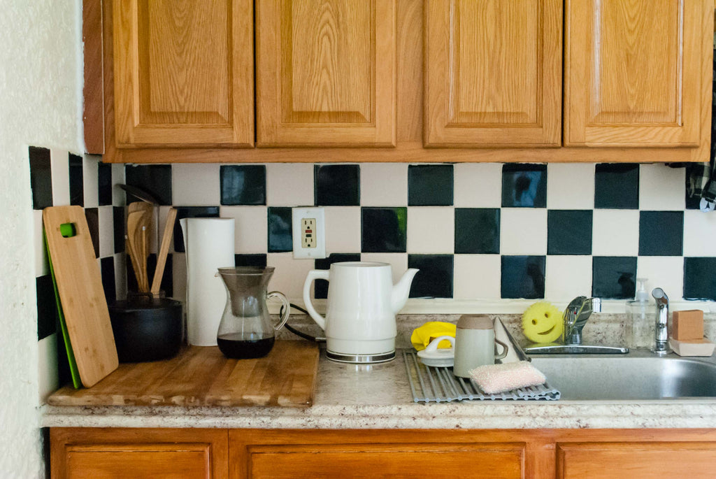 A small kitchen countertop with checkerboard backsplash, minimally accented with everyday items like an electric kettle and dish rack.