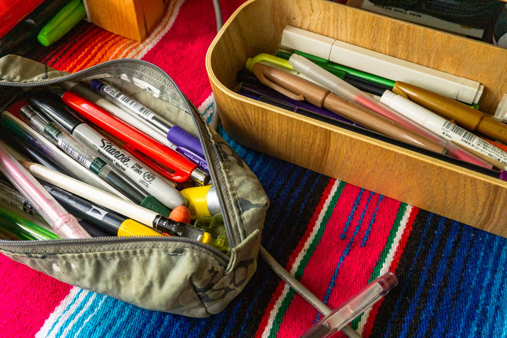 A cloth pencil case and a wood tray both full of pens and pencils on a colorful Mexican tablecloth.