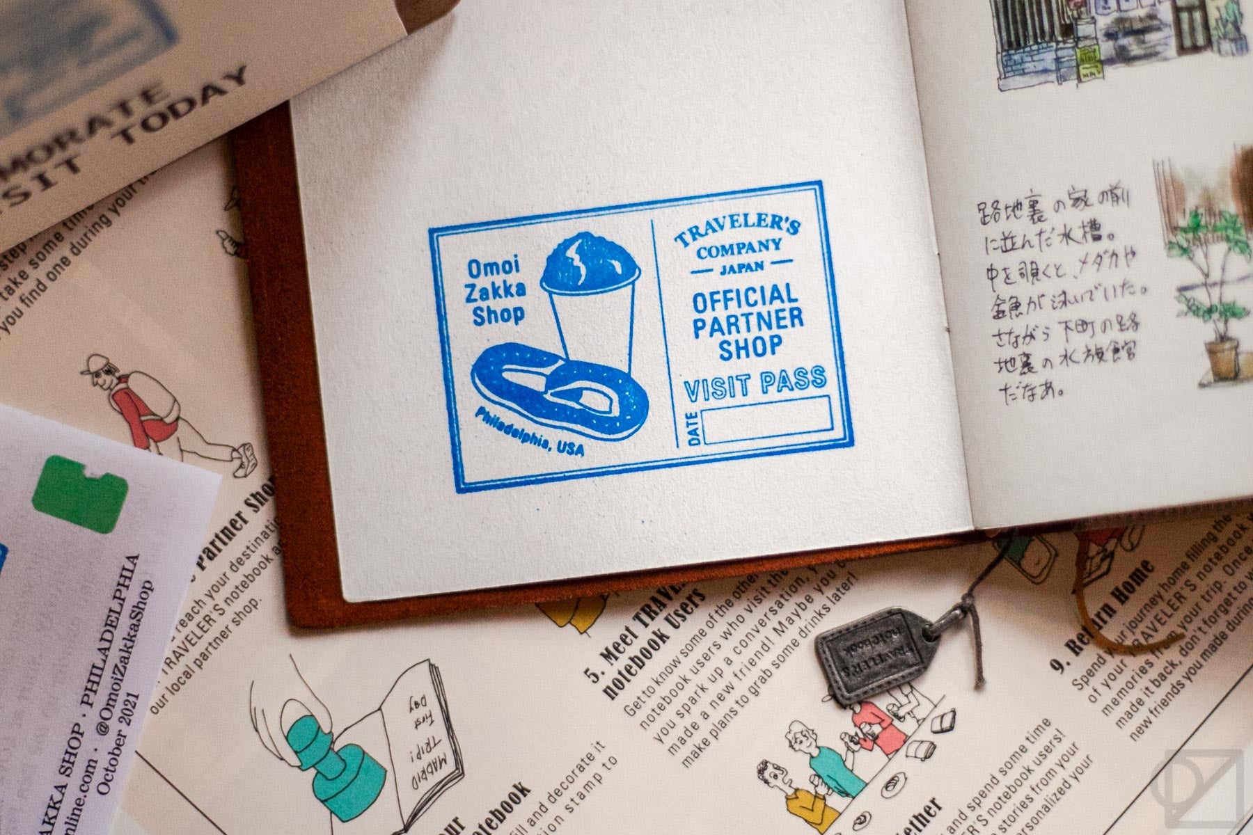 A close-up on the store stamp, blue ink.It says Omoi Zakka Shop with a drawing of a soft pretzel and water ice, and the official TRC logo, text saying visitor's pass, and place to write the date!