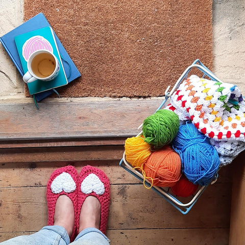 Feet wearing crochet slippers, stood on a doorstep with a basket of yarn and cup of tea