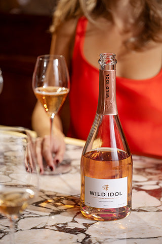A lady holding a glass of Wild Idol Alcolhol Free Sparkling Rosé Wine on table next to its bottle