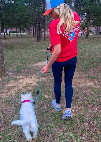 Woman in a red Rope Hounds shirt and Rope Hounds hat walking a white dog in a park.