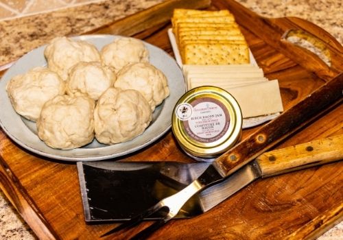 A wooden serving tray holds a plate full of bannock dough shaped into balls ready for roasting on an open fire. Next to the plate of dough is a jar of The Canadian Birch Company Birch Bacon Jam alongside crackers and cheddar cheese slices to be used to make Bacon Jam & Cheese S'mores on the fire as well.