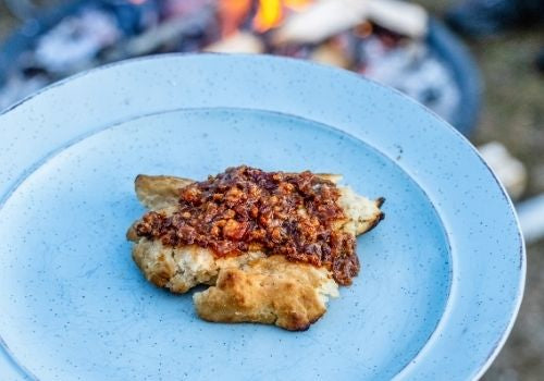 A thick piece of bannock rests on a robin's egg blue plate, with Birch Bacon Jam spread over the top, ready to be devoured.