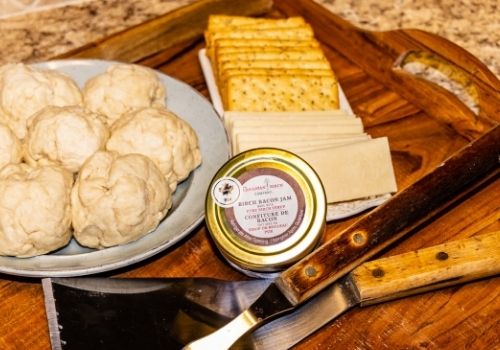A wooden carved tray with a plate of cheese and crackers, a fresh jar of birch bacon jam and bbq utensils in prep for making cheese and bacon jam s'mores over a camp fire. Also on the tray is a plate of bannock dough that can also be roasted on an open fire.
