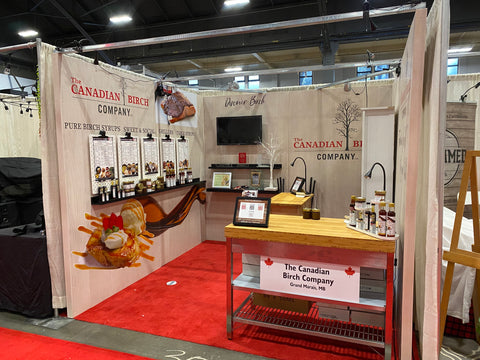 Photo of the newly designed Canadian Birch Company market booth with white washed wooden walls, food pairing posters mounted on sleek peg boards, TV screen with new birch themed videos and product display shelves.