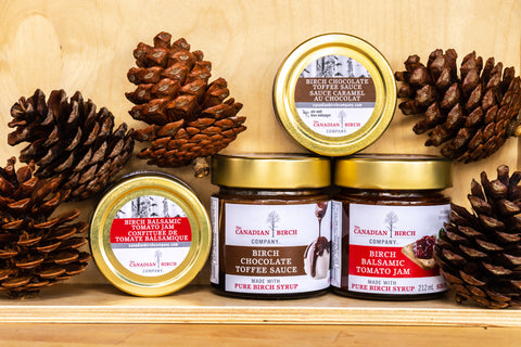 New Birch Balsamic Tomato Jam and Birch Chocolate Toffee Sauce in a stylized photo against a finished smooth birch wood background with the jars of product resting amongst large pine festive pinecones.