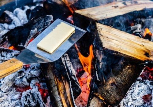 Over an open firepit fire with red coals and light flame, a metal BBQ spatula holds a thick slice of white aged cheddar as it begins to melt.