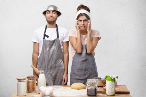 A man with a stainless steel baking bowl on his head and a woman holding her head in her hands are standing in the kitchen looking confused.