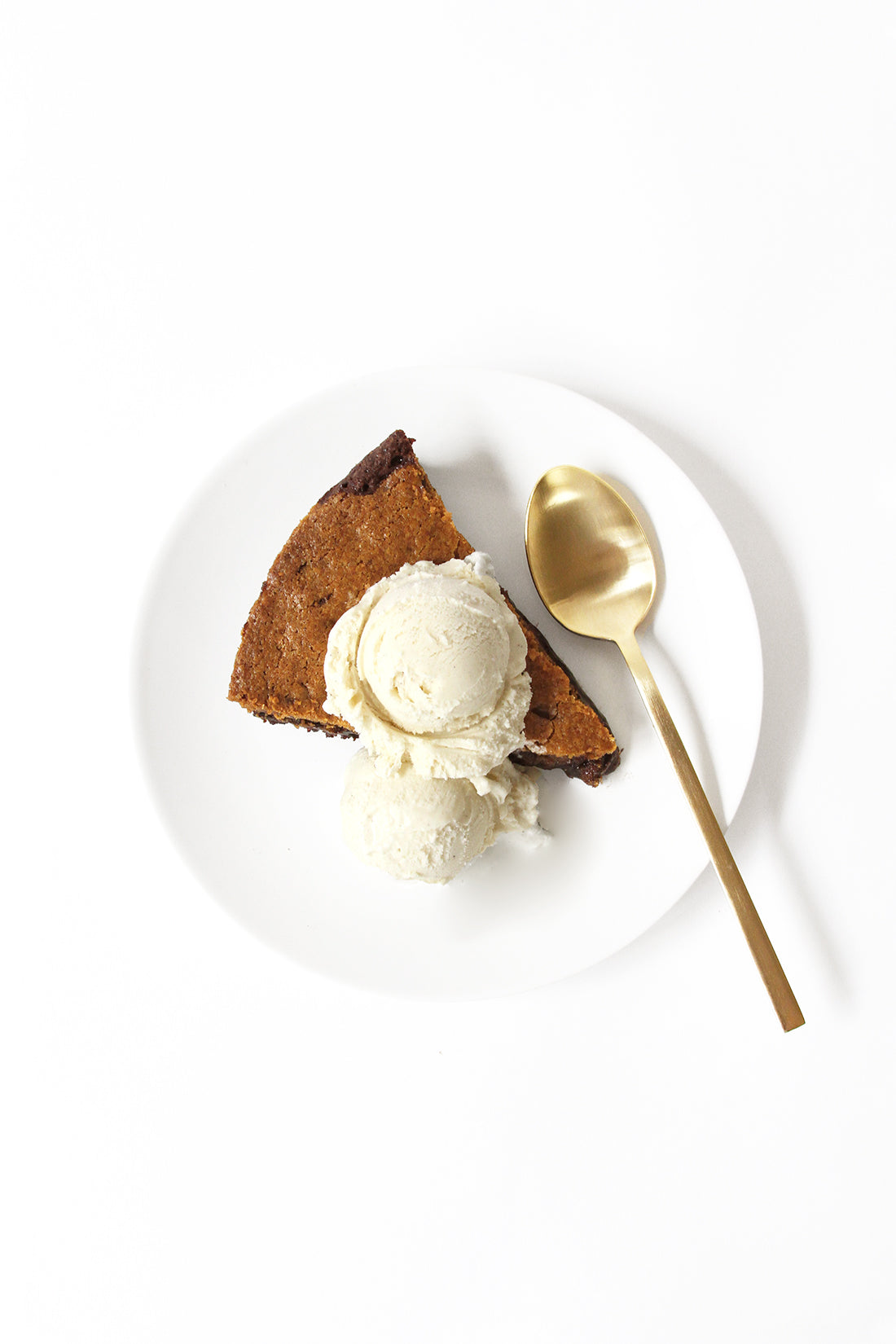 A slice of Miss Jones Baking Co Chocolate Chip Brownie Skillet on a white plate with a scoop of vanilla ice-cream on top next to spoon