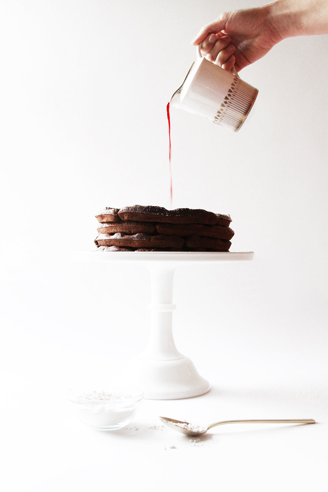 A hand pouring jam onto a stack of Miss Jones Baking Co Chocolate Cake Waffles on a cake stand