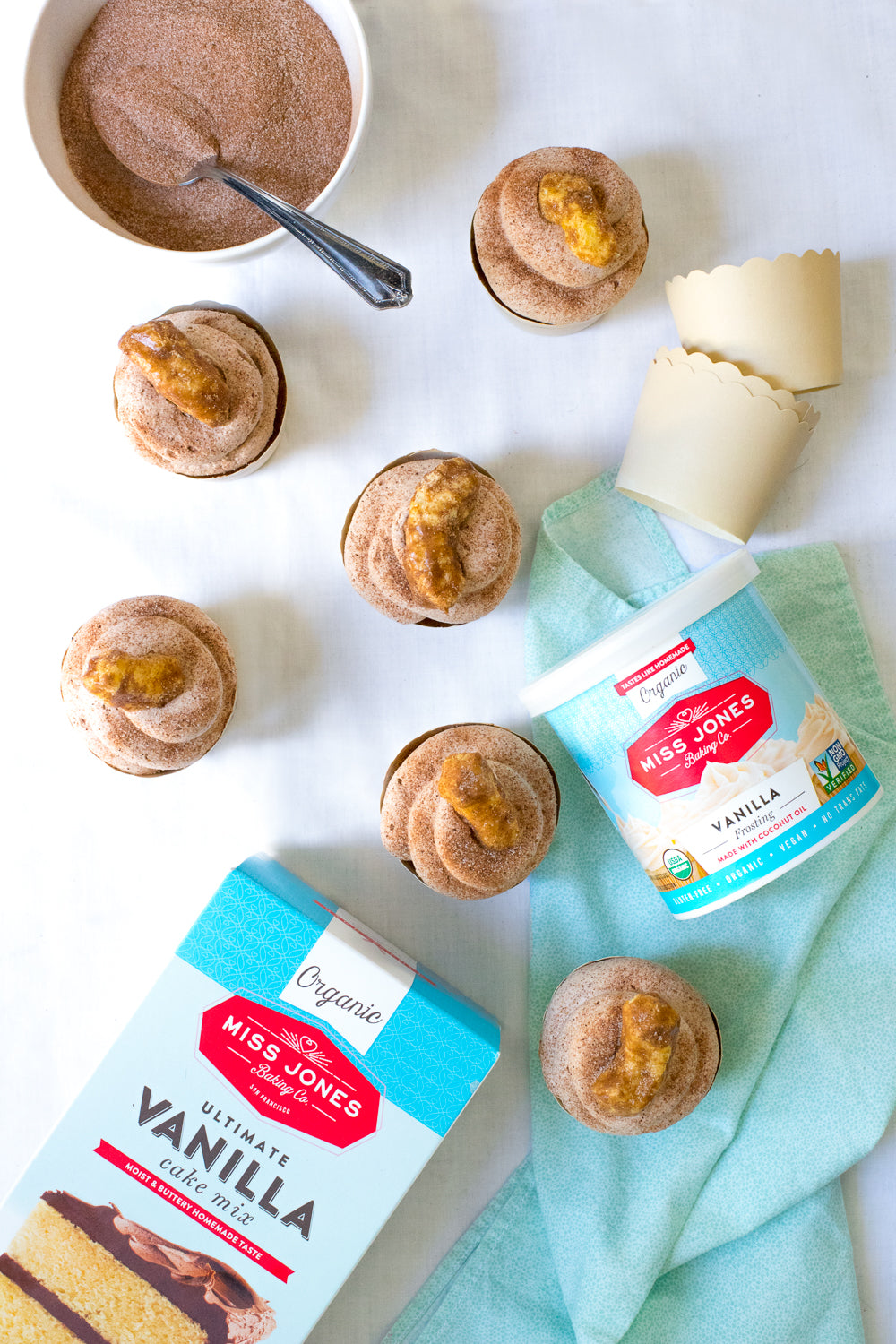 Image of six Miss Jones Baking Co Festive Churro Cupcakes next to a jar of Miss Jones Vanilla Frosting, a box of Miss Jones Vanilla Cake Mix and a bowl of cinnamon sugar