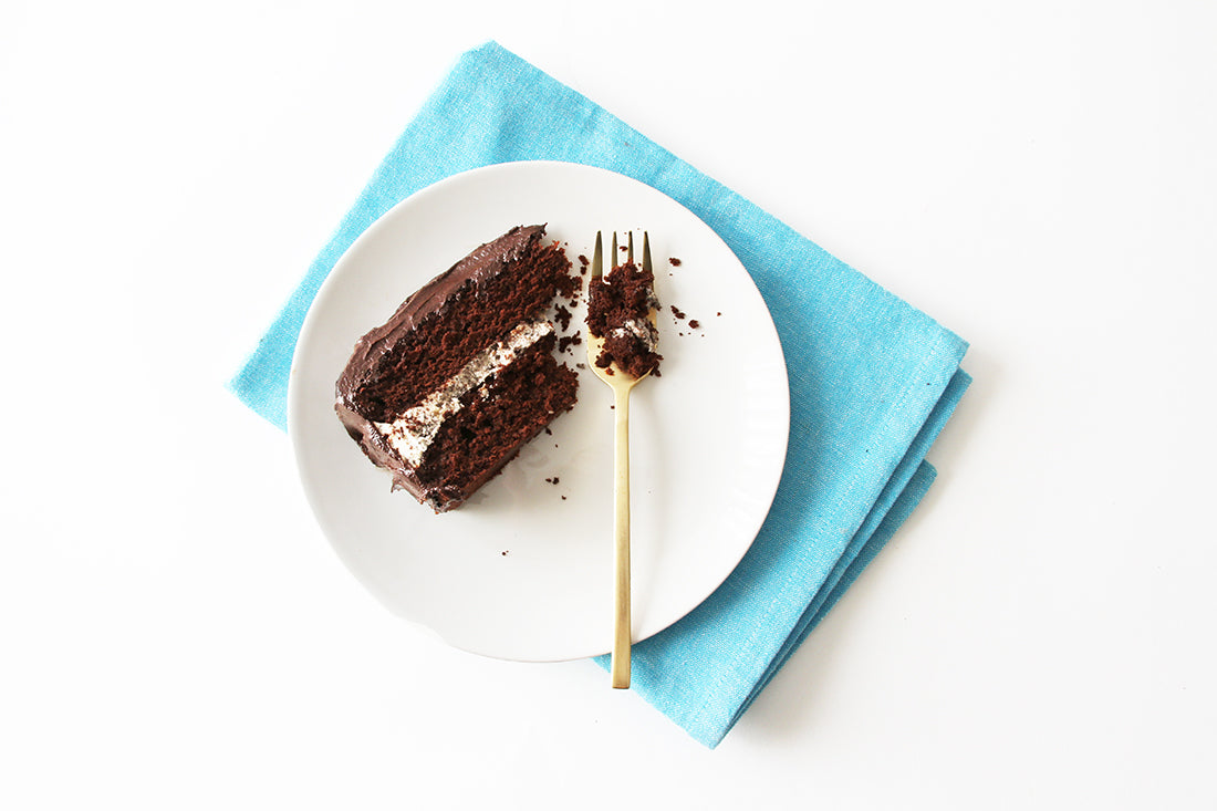 Image from above of a slice of Miss Jones Baking Co Minty Cookie Crunch Cake on a white plate next to a gold fork