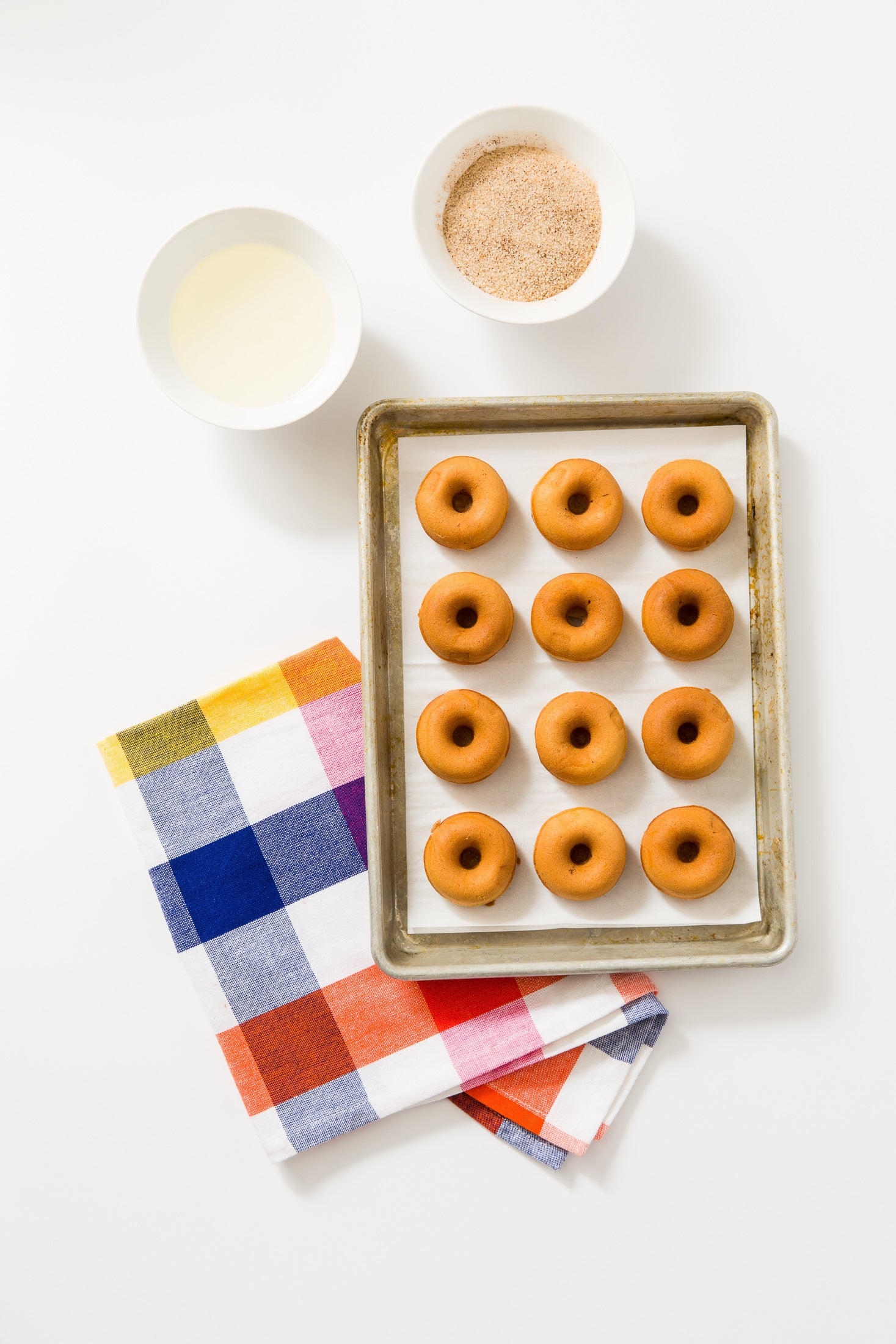 Image of twelve Miss Jones Baking Co Dirty Chai Mini Donuts on a baking sheet next to a bowl of milk and a bowl of cinnamon sugar