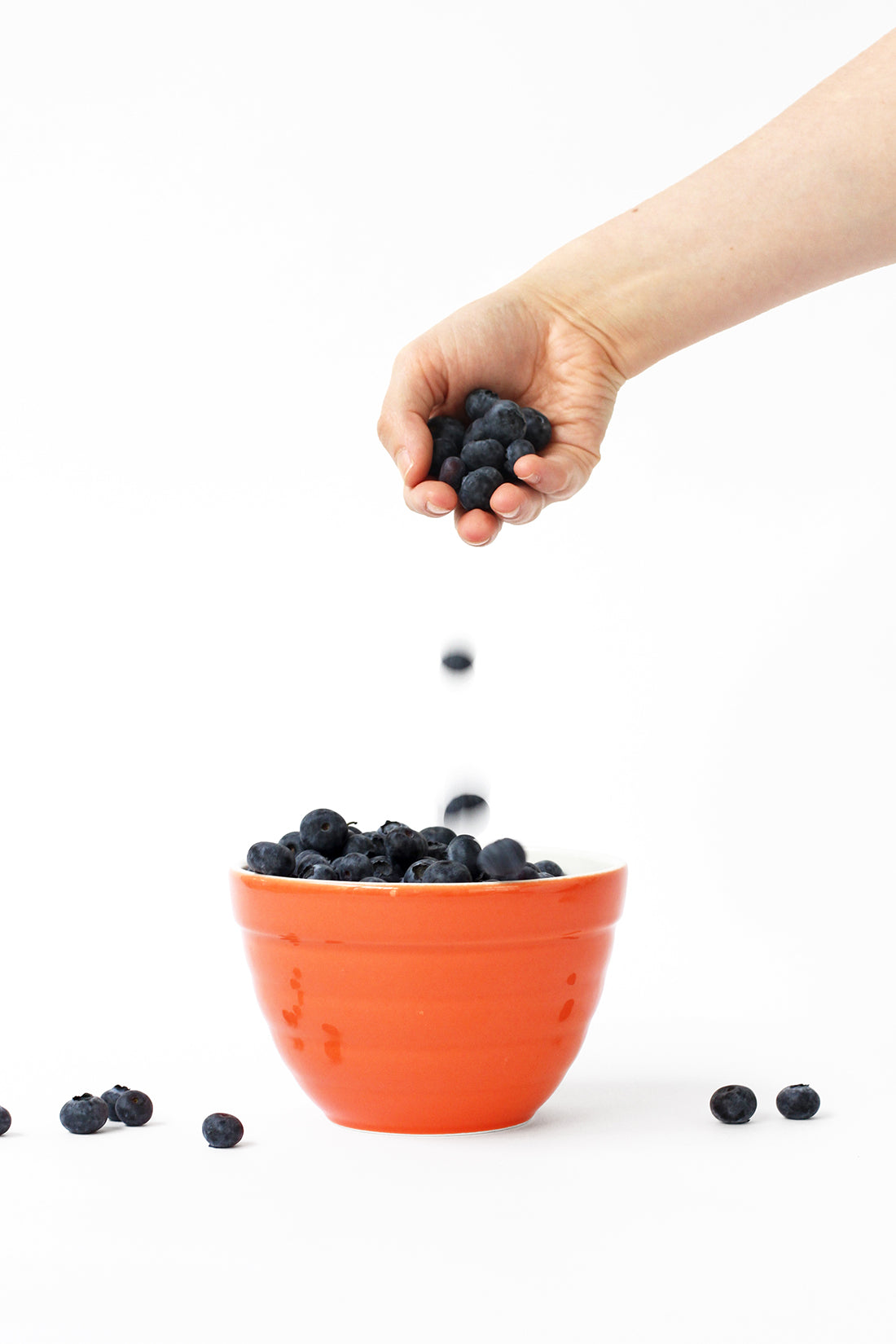 Image of a hand dropping blueberries into an orange bowl used for Miss Jones Baking Co Firecracker Pops recipe