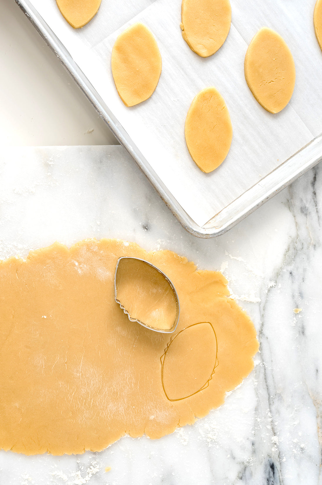 Image from above of rolled out cookie dough with a football cookie cutter next to football shaped cookie dough pieces on a baking rack for Miss Jones Baking Co Soft & Chewy Football Cutout Sugar Cookies