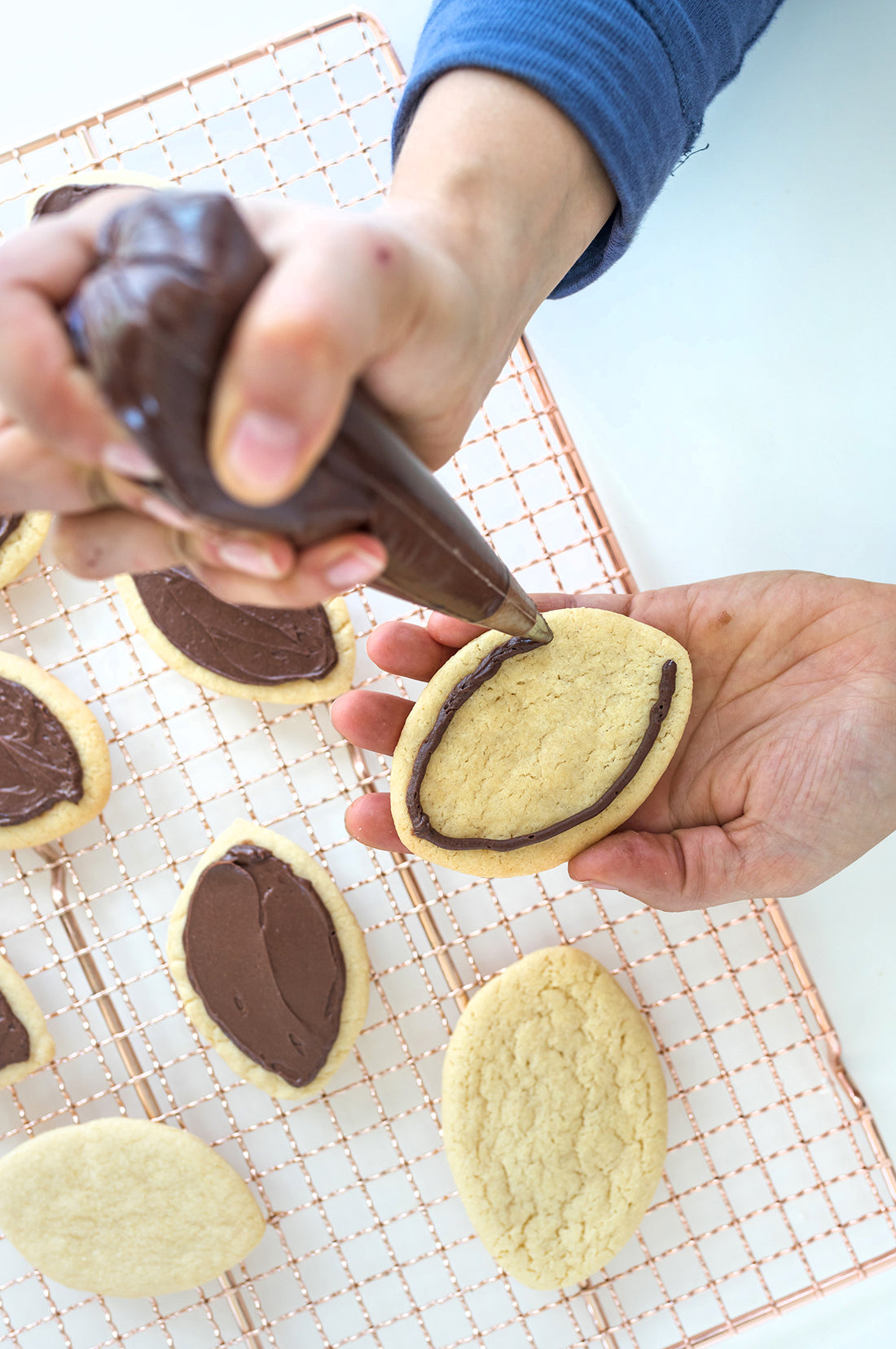 Image from above of football shaped sugar cookies on a baking rack being frosted with Miss Jones Chocolate Frosting for Miss Jones Baking Co Soft & Chewy Football Cutout Sugar Cookies