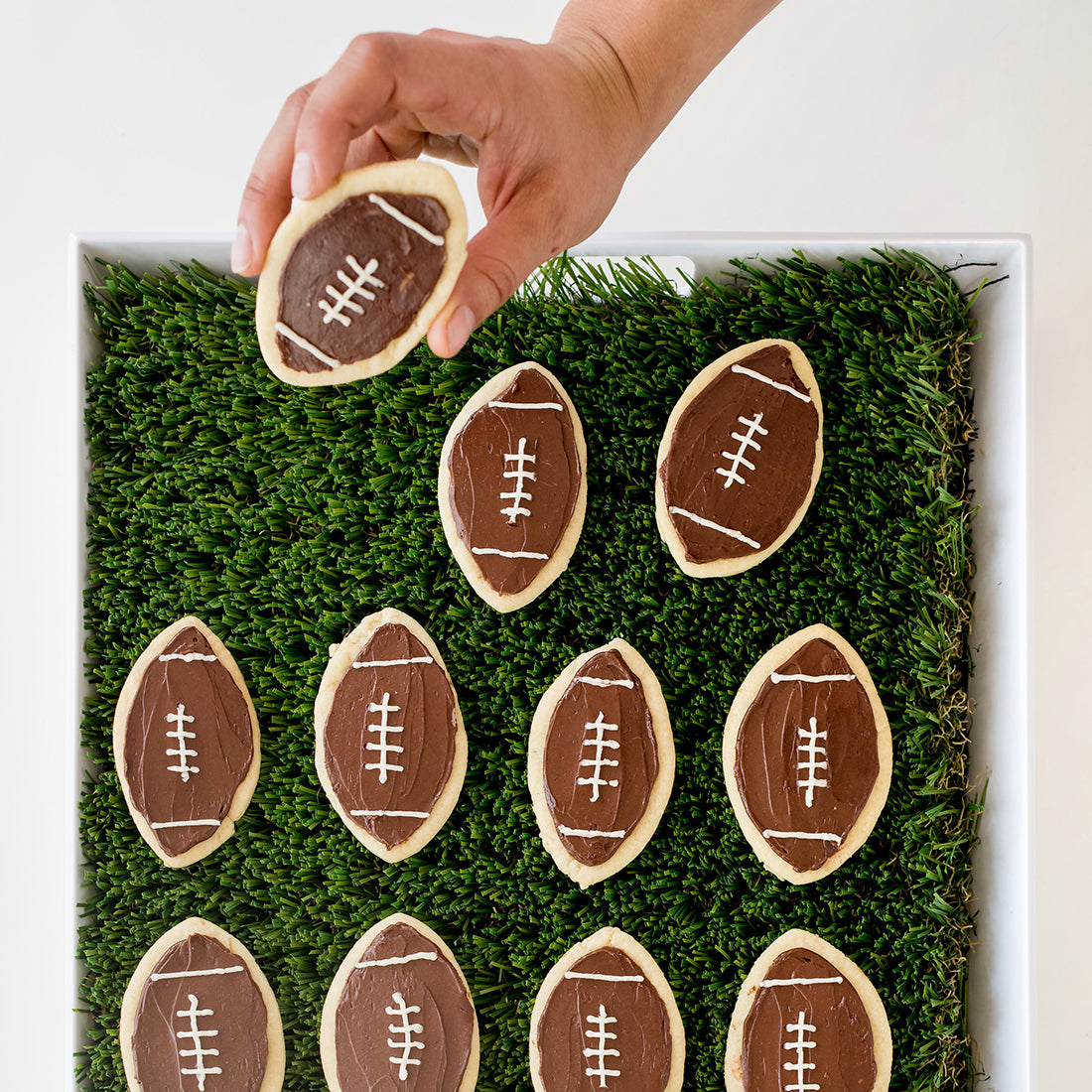 Image from above of eleven Miss Jones Baking Co Soft & Chewy Football Cutout Sugar Cookies on a turf mat