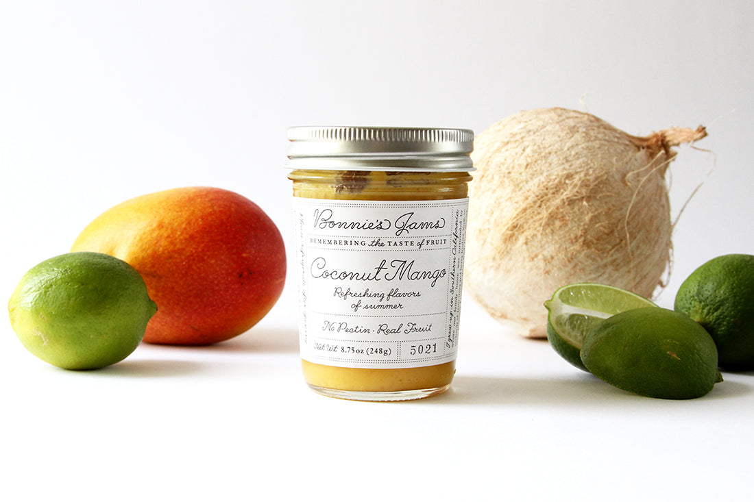 Image of side of a jar of Bonnie's Jams Coconut Mango flavored next to a coconut, a few limes, and a mango used for Miss Jones Baking Co Fruit Cart Tarts recipe