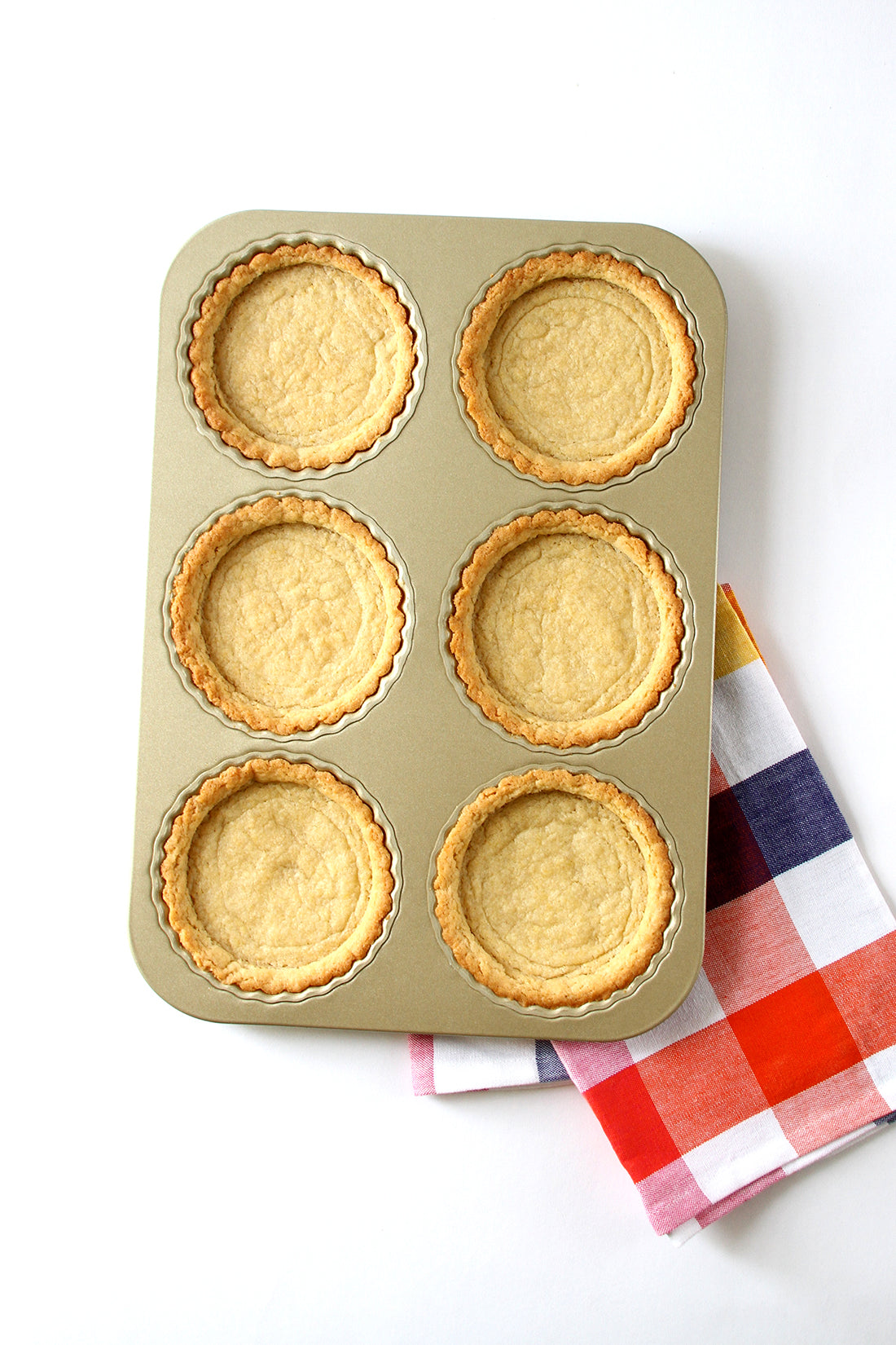 Image of six tart crusts in a baking pan on top of a checkered towel for Miss Jones Baking Co Fruit Cart Tarts recipe