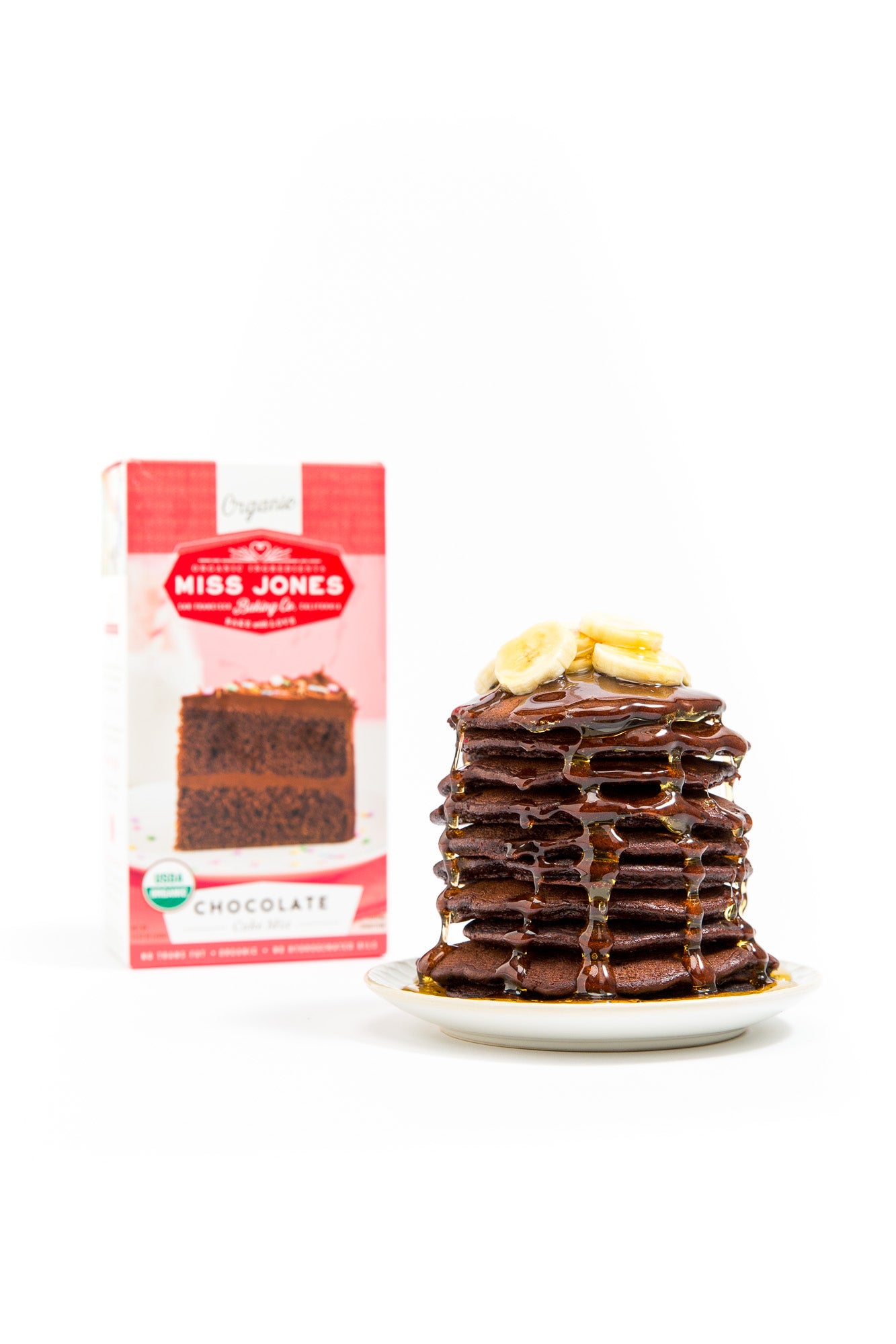 Stack of Miss Jones Baking Co Double Chocolate Stout Pancakes with melted butter and slices of banana on top with a box of Miss Jones Chocolate Cake Mix next to it