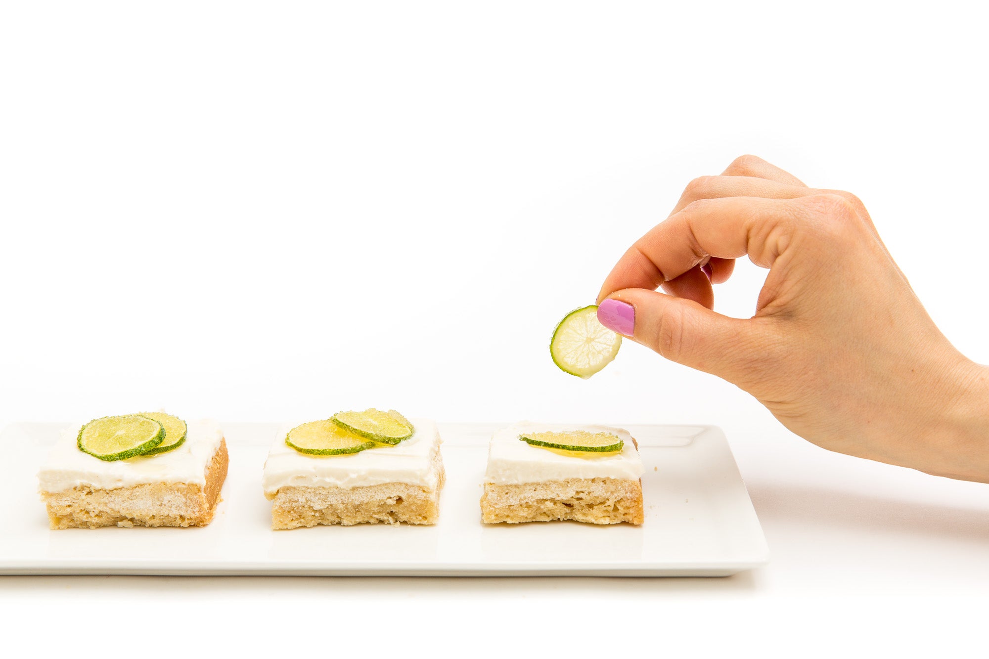 Image from the side of a hand putting baked lime slices on three Miss Jones Baking Co Key Lime Margarita Bars on a plate