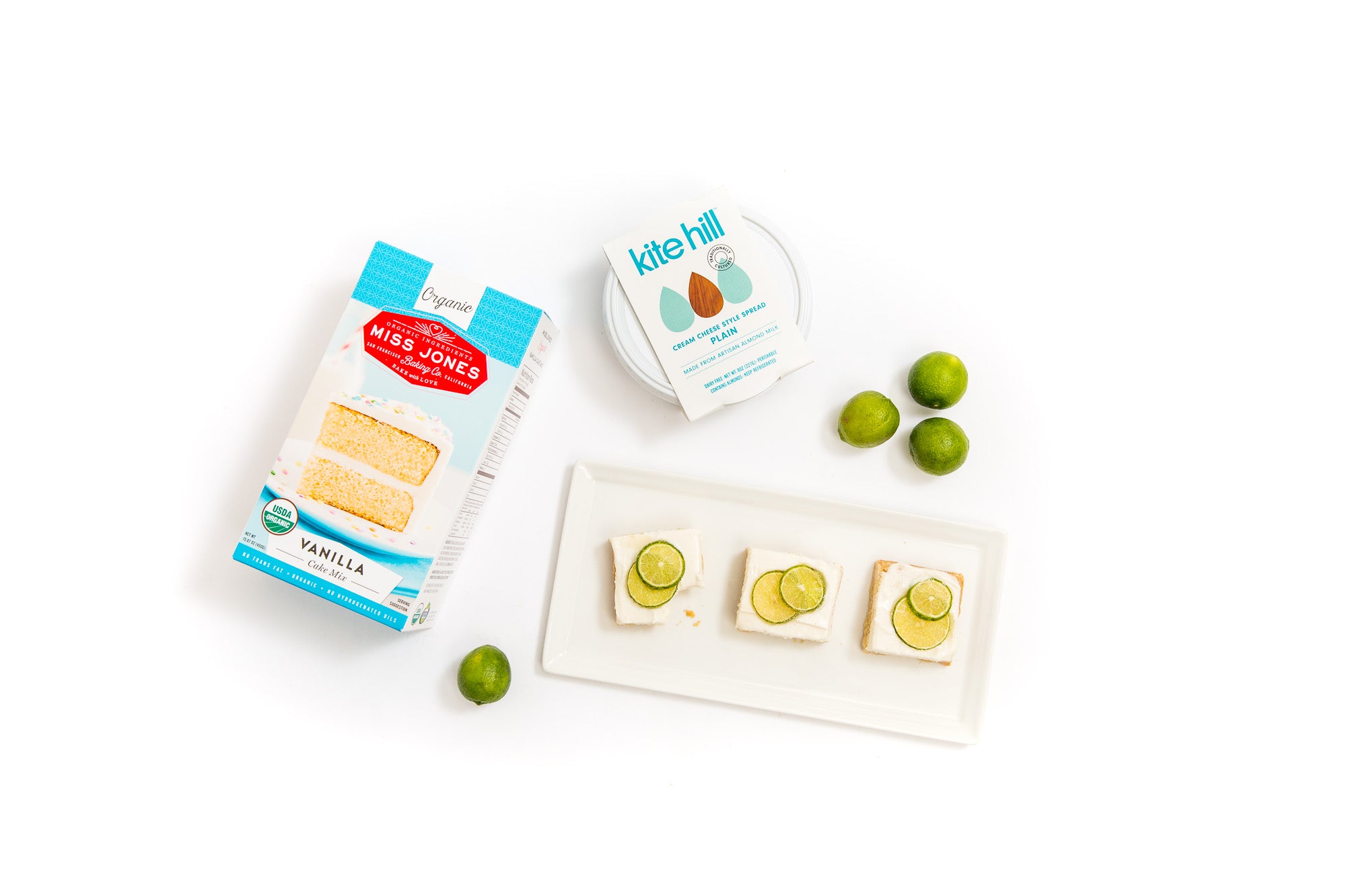 Image from above of a plate with three Miss Jones Baking Co Key Lime Margarita Bars, four limes, a box of Miss Jones Vanilla Cake Mix and a tub of Kite Hill Cream Cheese spread