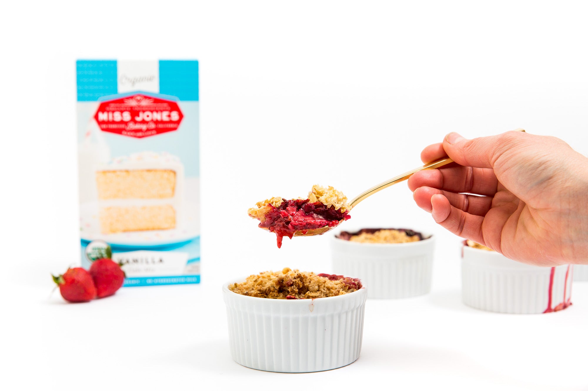 Image of a spoon holding a bite of a Miss Jones Baking Co Mini Berry Crumble in front of two more mini berry crumbles, a box of Miss Jones Vanilla Cake Mix and two strawberries