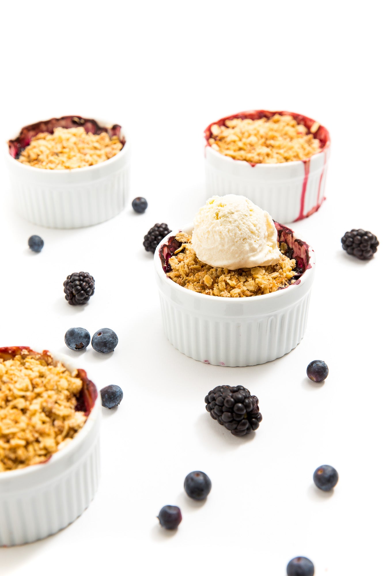 Image of four Miss Jones Baking Co Mini Berry Crumbles, one with a scoop of vanilla ice cream on top, surrounded by blackberries and blueberries