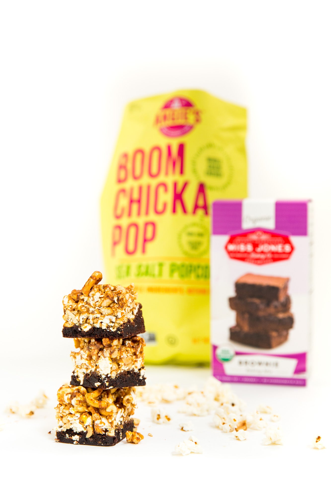 Image of three Miss Jones Baking Co Popcorn Brownies stacked in front of a box of Miss Jones Brownie Mix and a bag of Boom Chicka Pop Sea Salt Popcorn