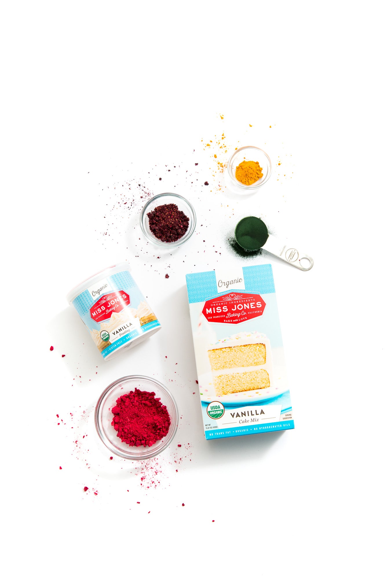 Image from above of a box of Miss Jones Vanilla Cake Mix, a jar of Miss Jones Vanilla Frosting and red, purple, green, and yellow food dyes