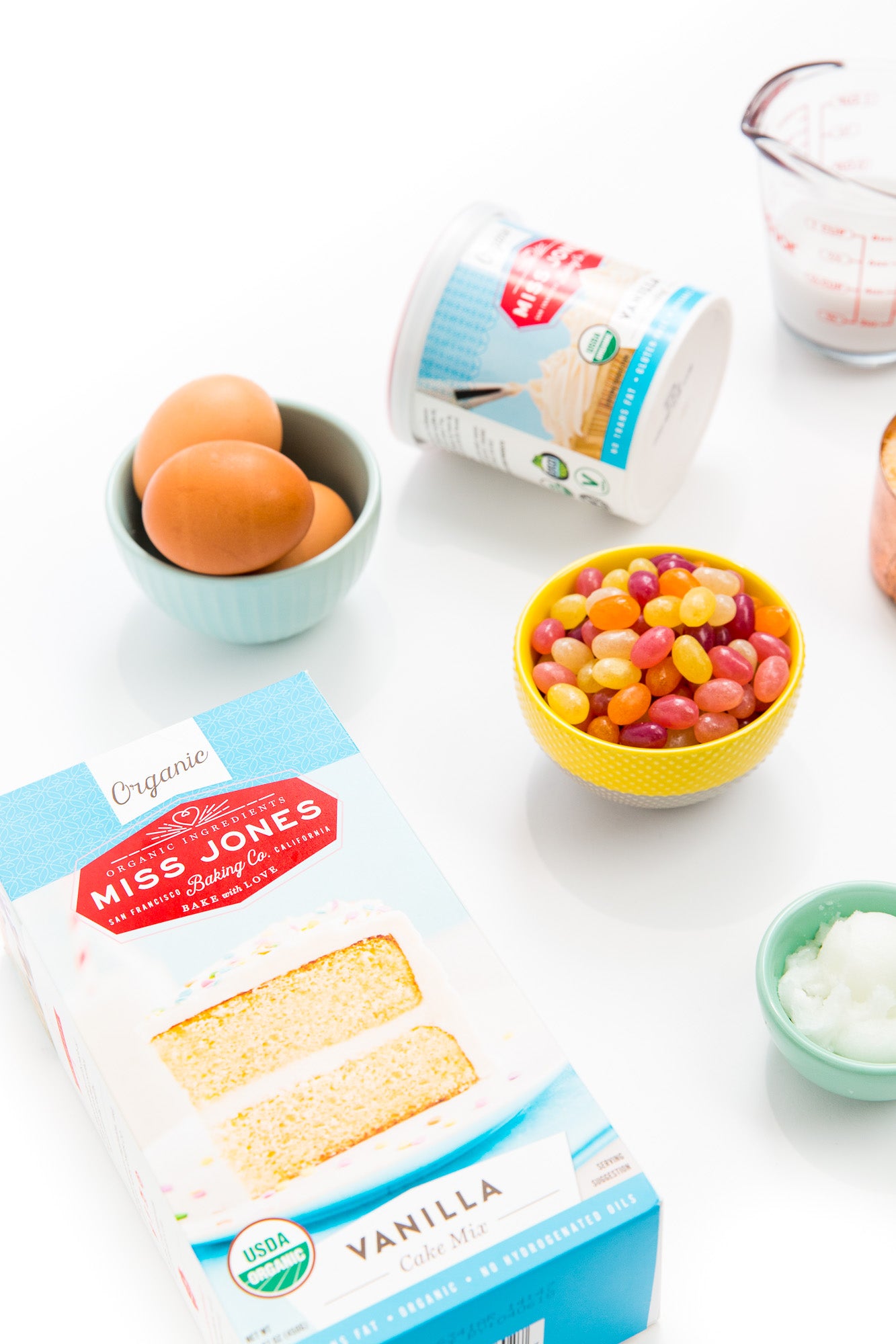 Image of a box of Miss Jones Vanilla Cake Mix, a bowl of three eggs, a bowl of jellybeans, a jar of Miss Jones Vanilla Frosting, and a bowl of coconut oil for Miss Jones Baking Co Toasted Coconut Easter Egg Nest Cupcakes recipe